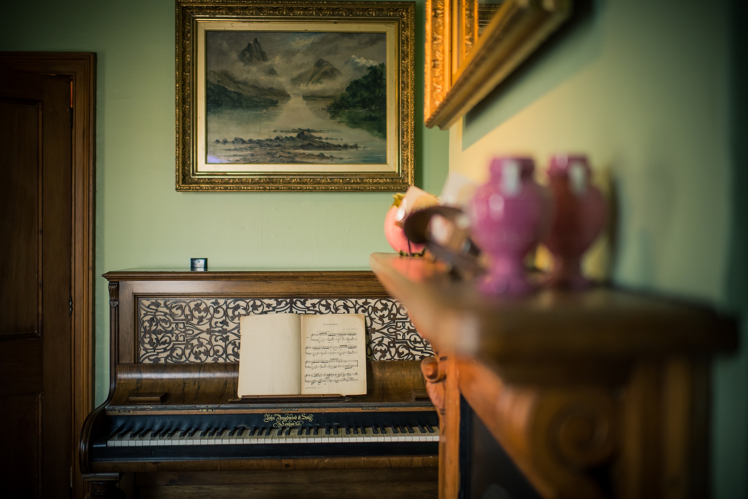 The pianola of the sitting room is against the pale green wall. The mantel piece in the foreground hold trinkets and is out of focus.