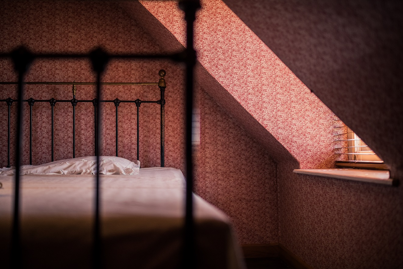 One of the cottage's bedrooms featuring soft pink. patterned wallpaper. Light comes through the window on to the made bed with white sheets and an iron frame.