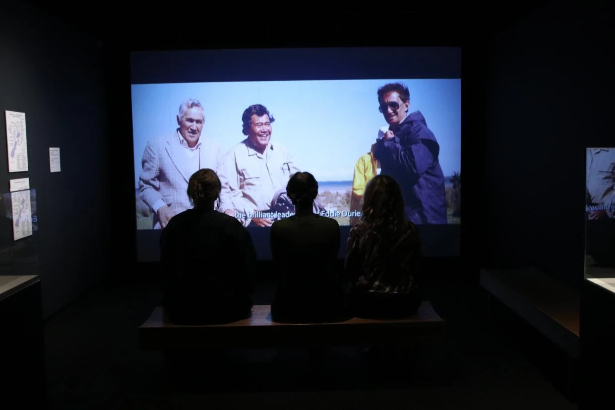 Three people are sitting on a bench in a dark room, watching a large screen that displays an image of three individuals standing outdoors. The room is part of an exhibition space with some framed items on the walls and another display case visible to the right.