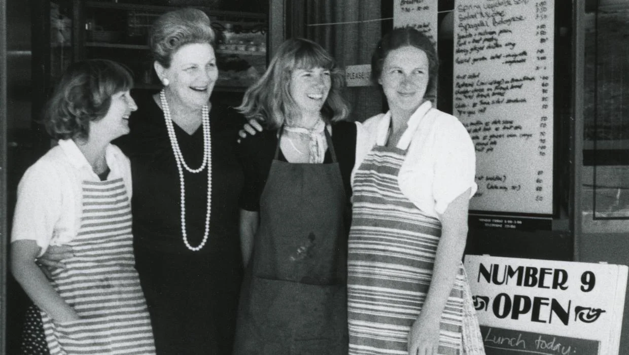 Four people stand in front of a restaurant or café. Three are wearing aprons, suggesting they may be staff. Behind them is a menu board listing food items and prices, along with a sign that reads 'NUMBER 9 OPEN' and 'Lunch today' underneath.
